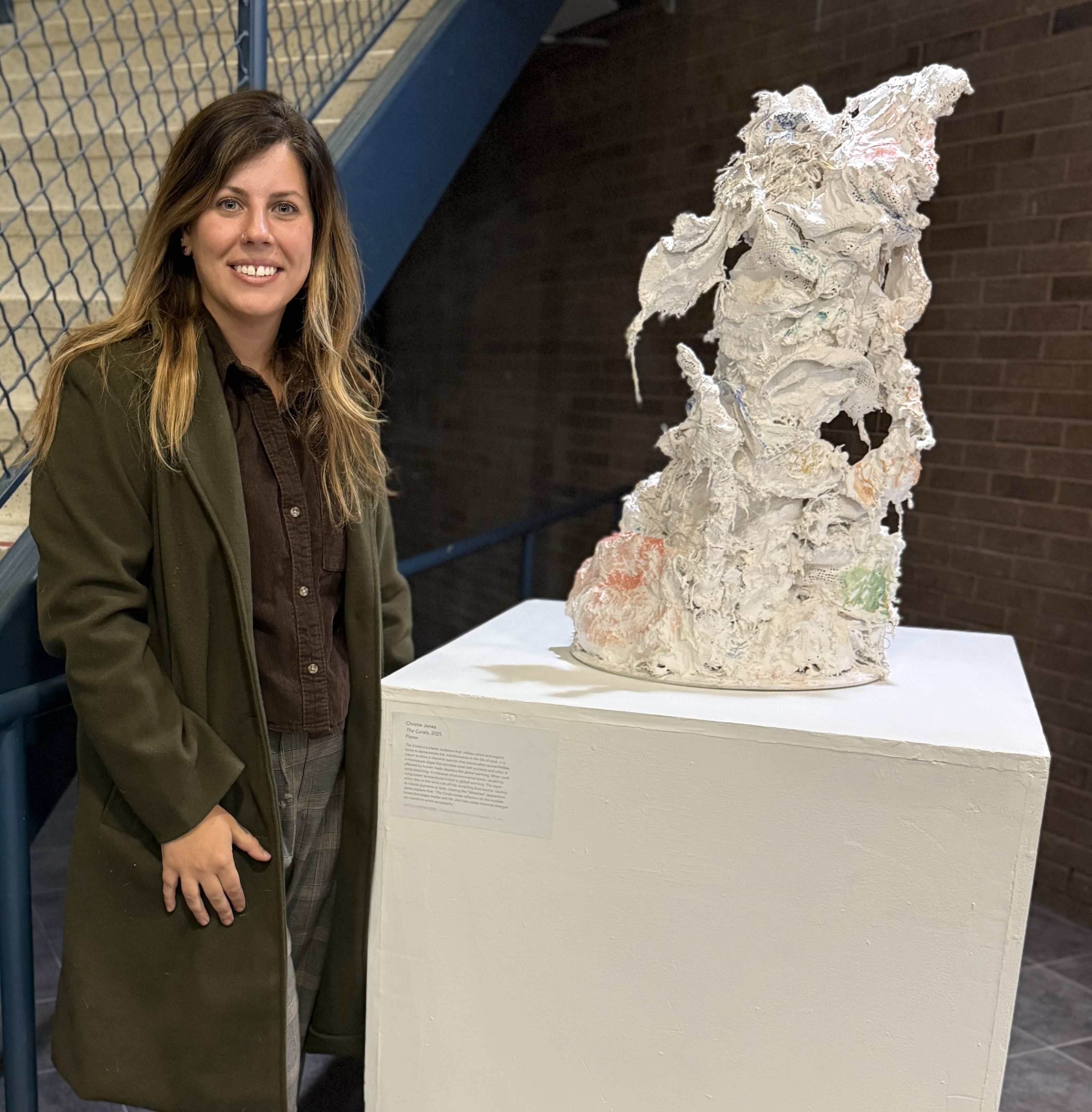 Christie Jones and Coral Sculpture Individual wearing a dark green coat stands next to a plaster sculpture on a white pedestal