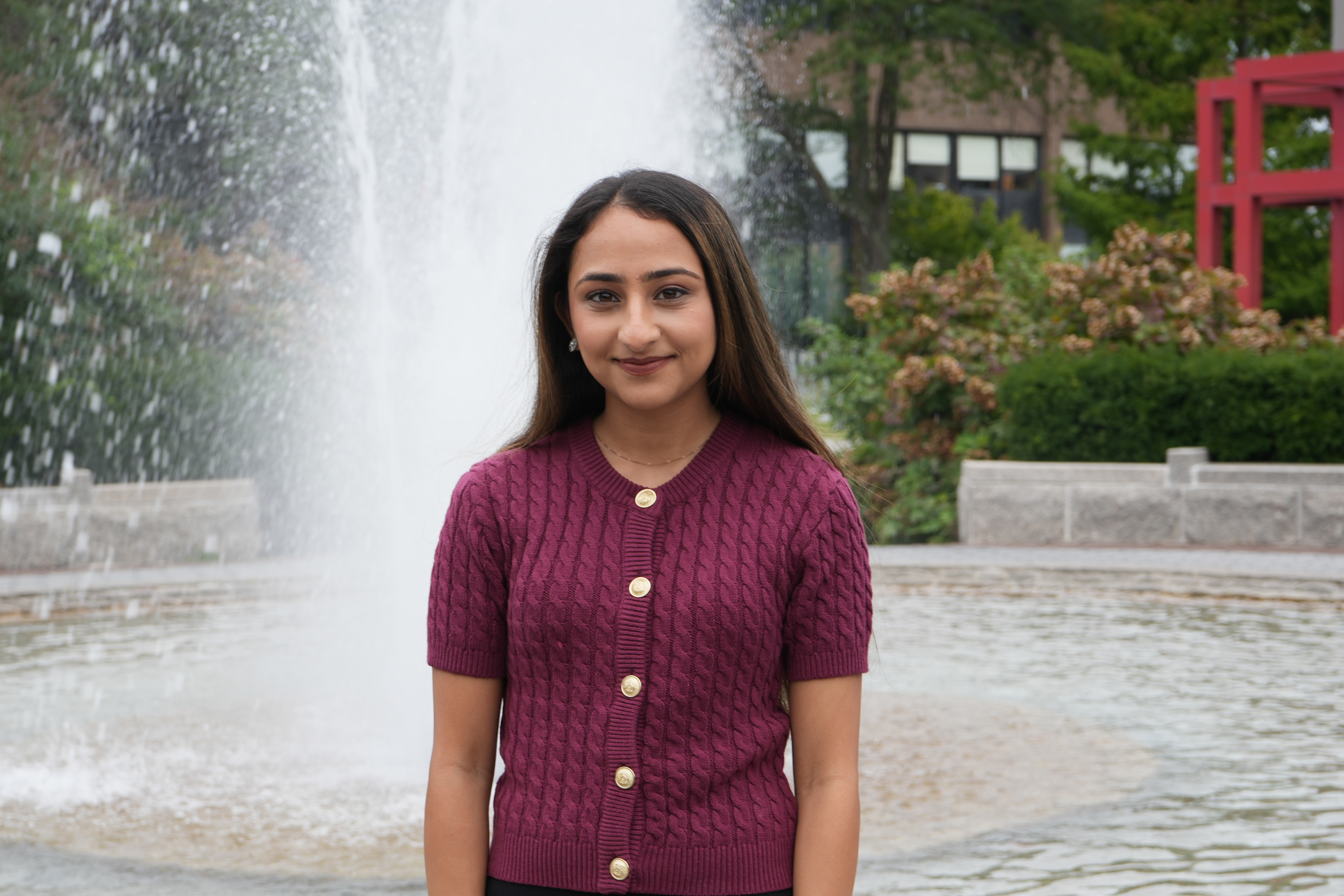 Laiba Rajput Person in a white button-up dress smiling, standing outside on a sunny day with greenery and a pathway in the background.