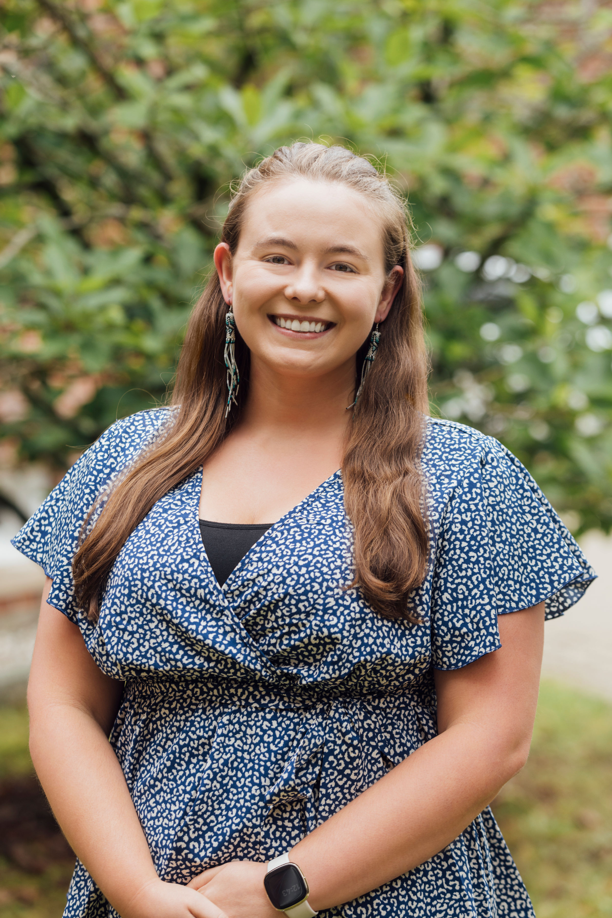 A person smiling at the camera, wearing a blue patterned dress, standing in front of a leafy background.