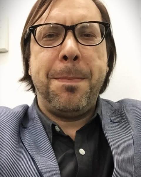 Close-up portrait of a person wearing glasses and a blue jacket over a dark shirt, smiling slightly at the camera.