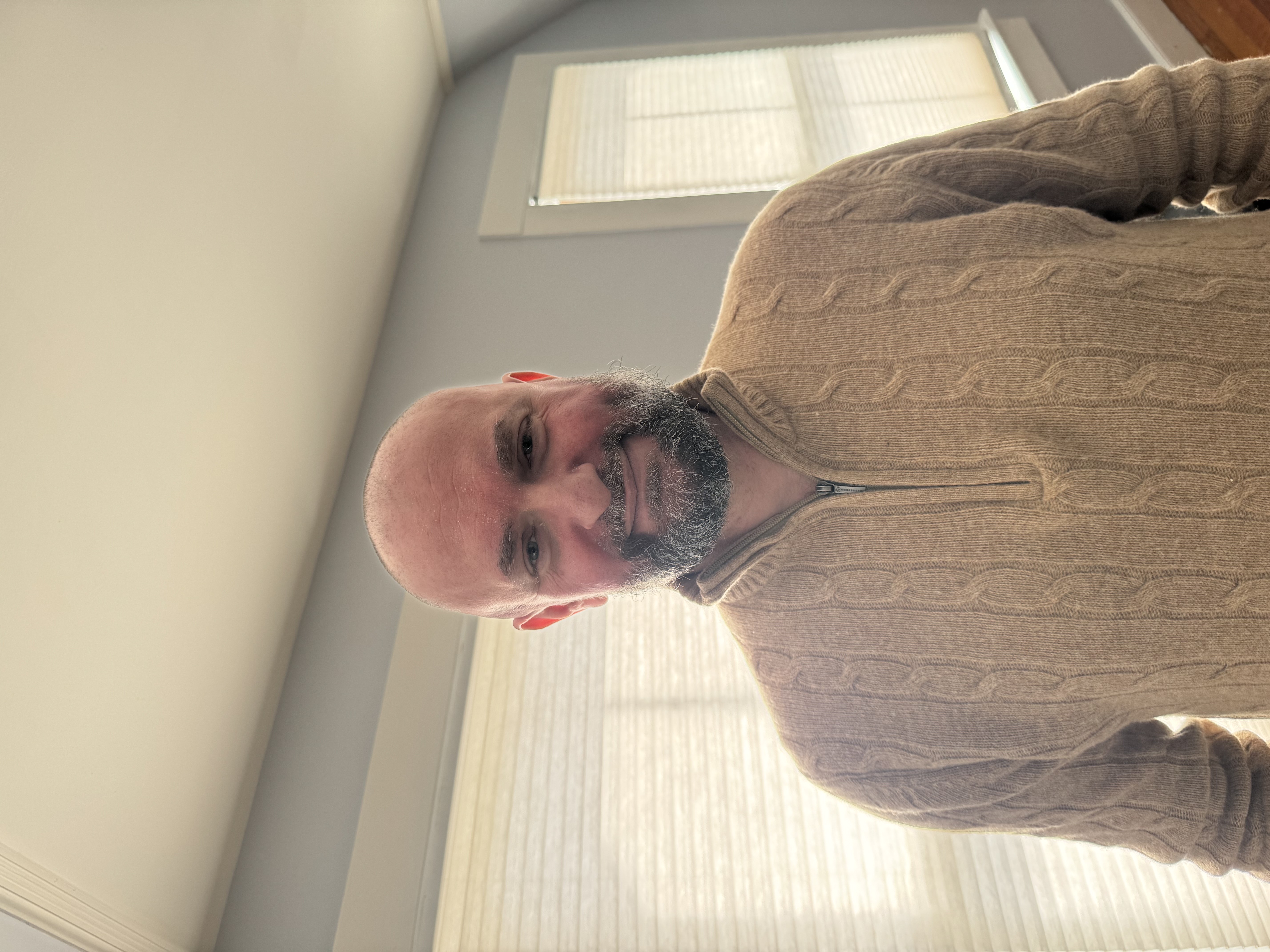 Person standing indoors wearing a beige sweater, with a beard and bald head, positioned in front of a window with white blinds diffusing bright light.