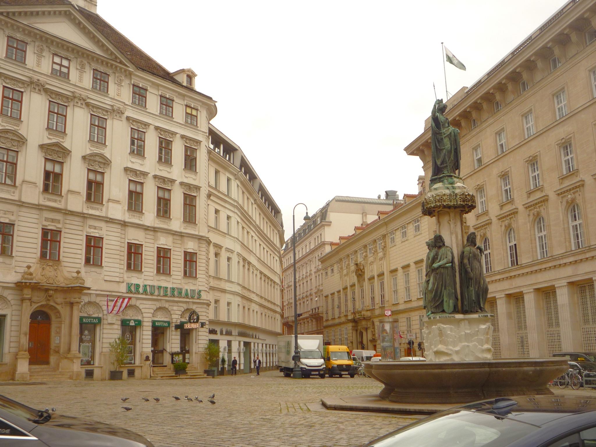 View of Judenplatz in Vienna, featuring the Lessing Monument, surrounded by classic European buildings and parked vehicles. Pigeons are scattered across the cobblestone square.