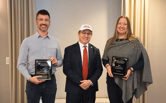 Excellence in Teaching Award Three people stand smiling in a room with neutral curtains. Two hold plaques, suggesting an award ceremony. The mood is celebratory and formal.