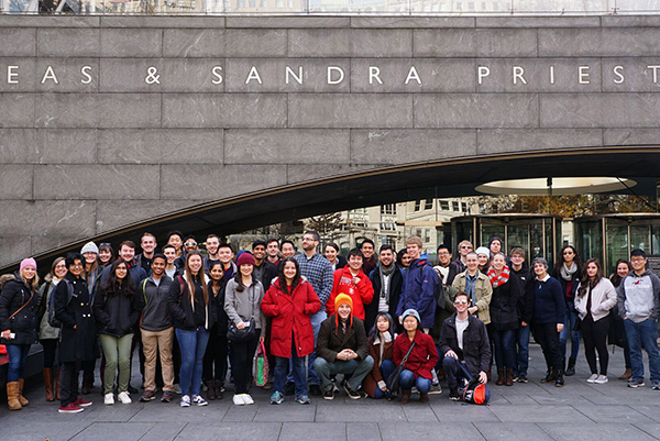 Honors College A group of people posing in front of a large building entrance, smiling and dressed in winter clothing. The mood is cheerful and the setting is urban.