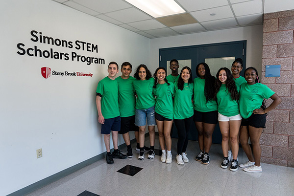Simons STEM Scholars A diverse group of ten smiling young adults wearing green shirts stands together in a hallway under a sign reading "Simons STEM Scholars Program" at Stony Brook University.