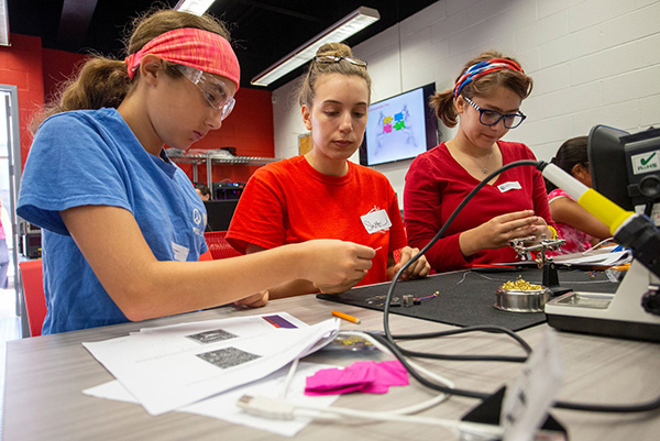 Women in Science (WISE) Honors Three young people in safety goggles and casual clothes work intently on electronics at a table in a workshop, reflecting concentration and teamwork.