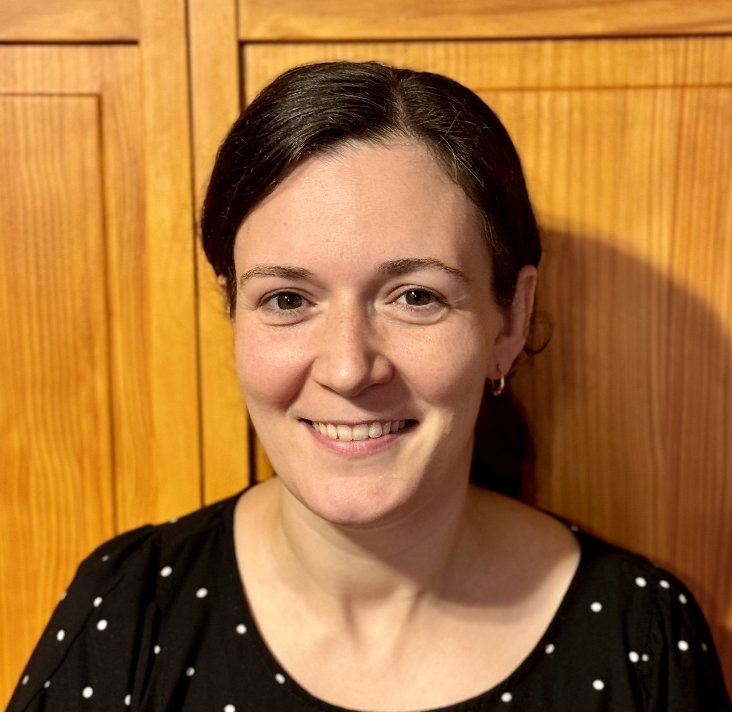 A portrait of a smiling woman with dark hair pulled back, wearing a black blouse with a white polka-dot pattern. She is centered in the frame against a background of warm-toned wooden cabinet doors.