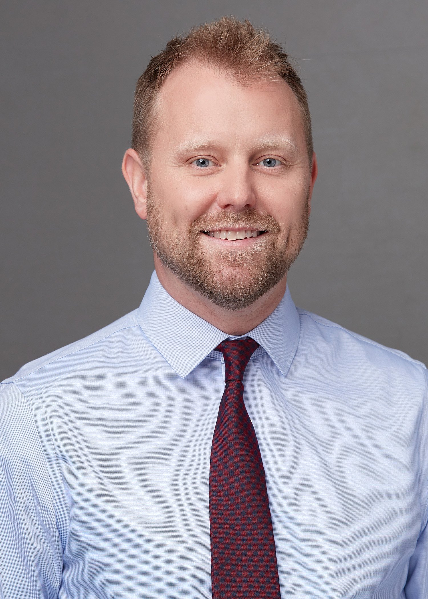 A professional headshot of a man with short, light brown hair and a groomed beard. He is smiling warmly at the camera, wearing a light blue dress shirt and a solid burgundy necktie. The background is a smooth, solid gray.