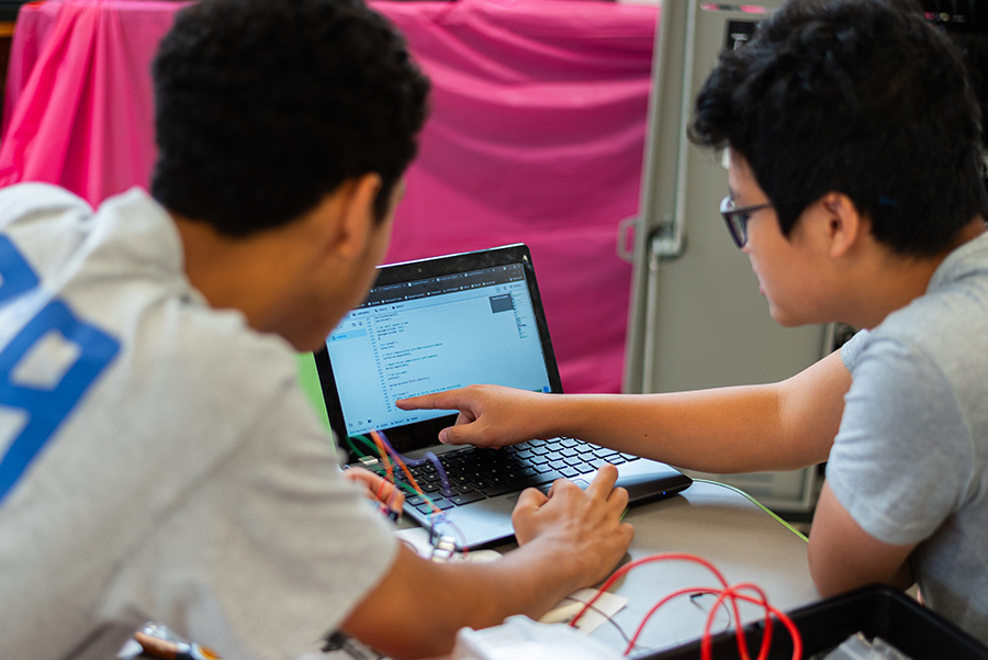 Two students looking at code on a laptop with parts of a robot on the desk in front of them