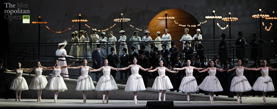 Dancers wearing white tutus on pointe in a line, in front of men and women dressed regally on a stage set of a street.